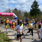 DSC_6023 Grupa biegaczy przekracza metę na świeżym powietrzu. Nad nimi różowy łuk z napisem META. Tło to drzewa i pochmurne niebo.