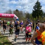 DSC_6028 Grupa biegaczy przekracza linię mety oznaczoną różowo-niebieskim łukiem, na tle zieleni i błękitnego nieba.