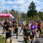 DSC_6033 Grupa biegaczy przekracza metę pod różowo-niebieskim balonem. Tło stanowi zielony las. Niebo jest częściowo pochmurne.