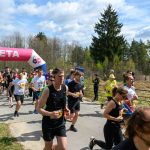 DSC_6036 Grupa biegaczy przekracza różowy baner z napisem META na leśnej ścieżce. Słoneczny dzień z niebieskim, chmurzastym niebem. W tle drzewa i kibice.