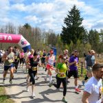 DSC_6037 Biegacze przekraczają różowo-białą metę na ścieżce w zielonym parku. Uczestnicy noszą kolorowe stroje sportowe. Słońce świeci, tworząc jasną i pogodną scenerię.