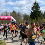 DSC_6041 Tłum ludzi biegnie w maratonie, przechodząc przez różowo-białą bramę z napisem META. Uczestnicy noszą kolorowe stroje sportowe. W tle widać drzewa i błękitne niebo z chmurami.