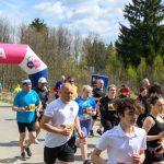 DSC_6049 Na zdjęciu widoczna jest grupa biegaczy przy mecie oznaczonej różowym balonem. Tło to zalesiony obszar i niebo z chmurami. Uczestnicy noszą kolorowe stroje sportowe.