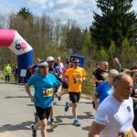 DSC_6050 Biegacze przekraczają różowy łuk z napisem META. W tle widać drzewa i niebo z chmurami. Dominują kolory niebieski i zielony.