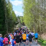 DSC_6055 Grupa biegaczy uczestniczy w wyścigu. Są ubrani w kolorowe stroje sportowe. Tło stanowi zalesiona droga i jasno-niebieskie niebo.
