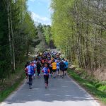 DSC_6057 Na zdjęciu grupa biegaczy na wąskiej drodze otoczonej zielonym lasem. Uczestnicy mają kolorowe stroje sportowe. Tło to jasne niebo i drzewa.