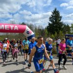 DSC_6069 Grupa ludzi kończy marsz nordic walking, przechodząc pod różowo-białym balonem z napisem META. Pogoda jest słoneczna z lekkimi chmurami, wokół widać zielone drzewa. Atmosfera jest żywa i sportowa.
