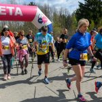 DSC_6071 Grupa ludzi w kolorowych strojach kończy wyścig nordic walking pod różowo-białą bramą mety. W tle drzewa i niebo. Z przodu kobieta w niebieskiej kurtce.
