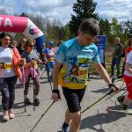 DSC_6072 Grupa ludzi maszeruje z kijami do nordic walking pod różowo-niebieskim banerem meta. Noszą kolorowe stroje sportowe. W tle drzewa i niebo z chmurami.