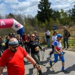 DSC_6074 Grupa ludzi z kijkami maszeruje pod różowym banerem oznaczonym META. Mają na sobie kolorowe ubrania sportowe. W tle widać drzewa i niebo z chmurami.
