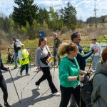 DSC_6081 Uczestnicy w sportowych strojach maszerują z kijami nordic walking. Tło stanowi zielone otoczenie z niebieskim niebem. W tle widoczny duży, biało-niebieski balon reklamowy.