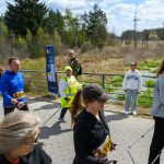 DSC_6083 Grupa ludzi uprawia nordic walking na chodniku. Tło to zielony krajobraz z drzewami. Niebo jest jasne i błękitne.