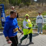 DSC_6084 Na zdjęciu widać dwie osoby nordic walking na tle lasu. Ubrani są na sportowo, jedna osoba w niebieskiej kurtce, druga w żółtej kamizelce. W tle są drzewa i młody chłopak w białej bluzie.