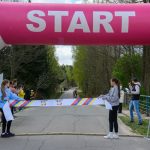 DSC_6091 Nad startem, różowy łuk z napisem START. Zawodnicy na drodze w lesie, osoby trzymają taśmę. Tło zielone drzewa.