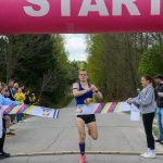 DSC_6097 Biegacz w niebieskiej koszulce przecina taśmę na mecie. Nad nim znajduje się różowy nadmuchiwany łuk z napisem START. W tle las i kibice.