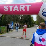 DSC_6109 Biegacz w czerwonym stroju przekracza linię startu pod różowym balonowym łukiem z napisem START. Tło stanowi asfaltowa droga otoczona drzewami. W tle widoczni są kibice i uczestnicy.