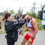 DSC_6113 Na zdjęciu kobieta wręcza medal mężczyźnie w stroju sportowym. Tło jest naturalne z drzewami, a wokół kibice. Widzimy również dmuchaną bramę mety.