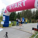 DSC_6119 Biegacz przekracza linię mety pod różowo-niebieskim łukiem z napisem START. Obok stoi zegar pokazujący 21:22. W tle widoczne drzewa i kilka osób.