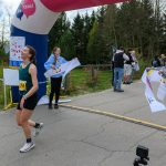 DSC_6128 Biegaczka przekracza linię mety, nosząc zieloną koszulkę i numer 74. Za nią łuk z napisem Ale czas!, a tło stanowi zieleń drzew. Dwie osoby trzymają taśmę mety, a obok stoi kilka osób.
