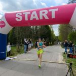 DSC_6134 Biegaczka w zielonych spodenkach przekracza linię startu pod różowym namiotem. W tle zielone drzewa i kibice. Po prawej cyfrowy zegar z czasem.
