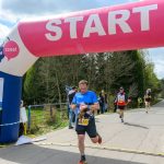 DSC_6136 Biegacze przekraczają linię startu mety z różowym napisem START. W tle drzewa i kibice przy drodze. Dzień słoneczny, niebo chmurne.