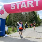 DSC_6138 Biegacze na starcie pod różowym łukiem z napisem START. Widoczni są kibice na tle zieleni. Obok łuku stoi tablica z mapą.