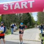 DSC_6139 Biegacze przekraczają różowy łuk startowy na wiejskiej drodze, otoczeni drzewami. Tablica czasowa wskazuje 25 minut. Na poboczu stoją gapie i organizatorzy.