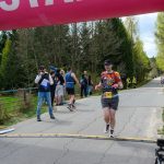 DSC_6140 Biegacz przekracza linię mety pod różowo-niebieskim łukiem START, otoczony drzewami i kibicami, czasomierz pokazuje 25 minut.