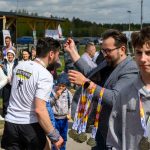DSC_6148 Zdjęcie przedstawia uczestników wydarzenia sportowego. Mężczyzna w okularach rozdaje medale ludziom w tłumie. Tło to zieleń i drewniany budynek.