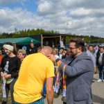 DSC_6154 Na zdjęciu mężczyzna w szarej marynarce zakłada medal osobie w żółtej koszulce. Tło to zielone drzewa i niebieskie niebo z chmurami. Wokół widać tłum ludzi, głównie ubranych casualowo.