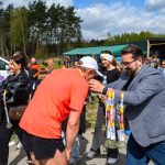 DSC_6156 Mężczyzna w szarej marynarce wręcza medale biegaczowi w pomarańczowej koszulce. W tle grupa ludzi i niebo z chmurami. Zieleń i namiot na dalszym planie.