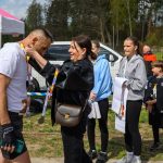 DSC_6158 Na zdjęciu kobieta wiesza medal na szyi mężczyzny. W tle namiot i grupa osób na zewnątrz, dzień jest słoneczny. Dominują kolory bieli, niebieskiego i zieleni.