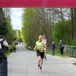 DSC_6161 Na zdjęciu biegnący uczestnicy maratonu na drodze otoczonej drzewami. Kobieta w zielonej koszulce uśmiecha się, mijając linię mety. W tle widoczni kibice i zieleń.