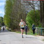DSC_6168 Na zdjęciu biegacz w zielonej koszulce biegnie po asfaltowej drodze w lesie. Po bokach trasy stoją widzowie. W tle widać drzewa i zachmurzone niebo.