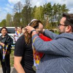 DSC_6171 Osoba wręcza medale uczestnikom na zewnątrz. Kolorowe wstążki, ludzie w tle, zielone drzewa. Słoneczny dzień.