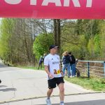 DSC_6174 Biegacz przekracza linię mety pod różowym bannerem startowym. Tło z zielonymi drzewami i kilkoma widzami. Zegar pokazuje czas 28:07.