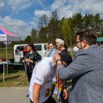 DSC_6176 Mężczyzna wręcza medale zawodnikowi na zawodach na tle namiotu. Dookoła stoją widzowie. Niebo jest częściowo zachmurzone.