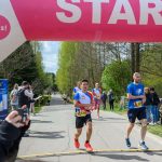DSC_6181 Biegacze startują pod różowo-białym łukiem z napisem START. W tle zielone drzewa i niebo. Obok kibice z aparatami.