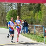 DSC_6183 Dwóch biegaczy przekracza linię mety podczas zawodów. Mają na sobie kolorowe stroje sportowe, a tło to zielone drzewa. Obok widać licznik czasu, pokazujący 28 minut 45 sekund.