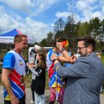 DSC_6186 Biegacze w kolorowych koszulkach przy namiocie na tle lasu. Słoneczna pogoda, ludzie wokół robią zdjęcia. Rozdawanie medali.