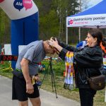 DSC_6192 Na zdjęciu kobieta zakłada medal mężczyźnie na mecie biegu. Tło to zielone drzewa i namiot. Widoczna tablica z czasem 29:19.