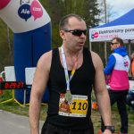 DSC_6196 Zawodnik w biegu, numer 110, ubrany na czarno, z medalem na szyi. Tło to zielone drzewa i niebieski dmuchany łuk z napisem. Po lewej licznik czasu, z prawej biały namiot z reklamą.