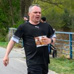 DSC_6203 Mężczyzna biegnie w maratonie, ubrany na czarno z białymi akcentami, trzymając numer startowy. Tło z zielonymi drzewami i niebieską barierką. Zdjęcie zrobione na zewnątrz, podczas cieplejszego dnia.