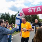 DSC_6205 Na zdjęciu mężczyźni kończą bieg pod różowym łukiem startu i mety. Pierwszy z nich ma pomarańczową koszulkę, obok są kibice. W tle widać drzewa i niebieskie niebo.