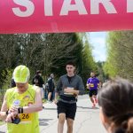DSC_6218 Biegacze na trasie pod różowym banerem START. Dzień słoneczny, drzewa w tle, kibice po bokach dopingują.