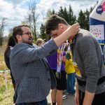 DSC_6221 Mężczyzna zakłada medal na szyję uczestnika biegu. Tło stanowi zielone pole i drzewa pod niebieskim niebem. Wokół widać innych uczestników.