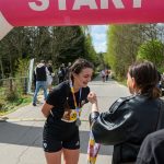 DSC_6234 Zawodniczka w czarnej koszulce otrzymuje medal pod różowym łukiem startowym. W tle zieleń i kilku biegaczy. Ciepły, słoneczny dzień.