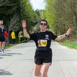 DSC_6238 Biegaczka przekracza linię mety na leśnej drodze. Ubrana na czarno, w tle zielone drzewa. Na koszulce numer 18.