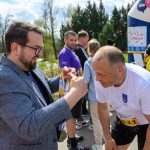 DSC_6246 Na zdjęciu mężczyzna w okularach wręcza medal biegaczowi w białej koszulce na tle zieleni. W tle kilka osób i dmuchany łuk. Kolory są jasne, przeważają niebieski, biały i zielony.
