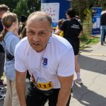 DSC_6247 Biegacz w białej koszulce odpoczywa po biegu. W tle widać balon z napisem „Ale czas!” i ludzi. Tło jest zielone, z drzewami.